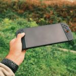 Close-up of a person's hand holding a black handheld gaming console against a green outdoor background. – China's mobile gaming dominance