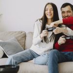 Joyful female and male in casual clothes sitting on comfortable sofa in living room while laughing and playing video game – Epic Games Store free