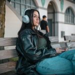 A woman in a leather jacket listens to music on a city bench in Vancouver. – JBL Live 780NC and 680NC Noise-Cancelling Headphones