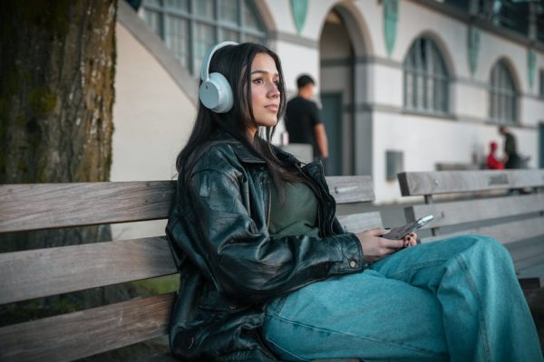 A woman in a leather jacket listens to music on a city bench in Vancouver. – JBL Live 780NC and 680NC Noise-Cancelling Headphones