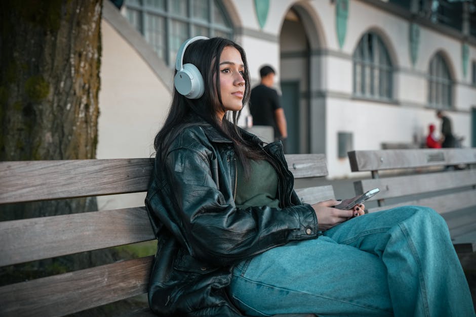 A woman in a leather jacket listens to music on a city bench in Vancouver. – JBL Live 780NC and 680NC Noise-Cancelling Headphones