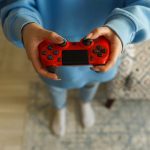Person holding a bright red gaming controller indoors, focusing on gameplay. – mass layoffs in video games