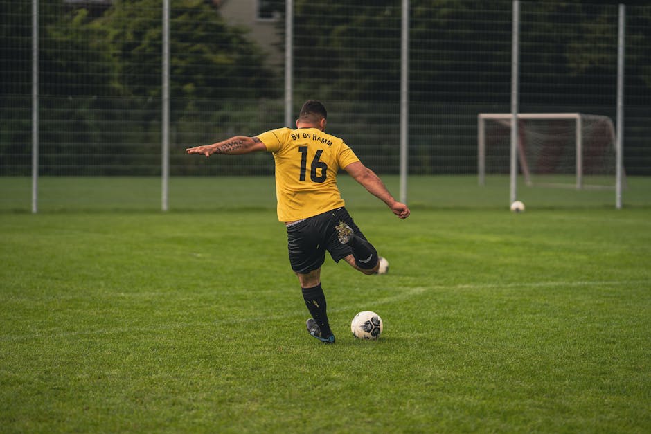 Back view of a football player in yellow jersey kicking a ball on a grass field.